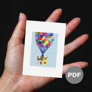 Puede incluir: Un patrón de punto de cruz que representa una casa con una fachada amarilla y un techo rojo, levantada por un grupo de globos de colores.