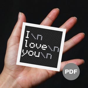 May include: A small, square cross-stitch design with the words "I love you" in white letters on a black background. The design is held in a hand against a dark background. A PDF icon is in the lower right corner.