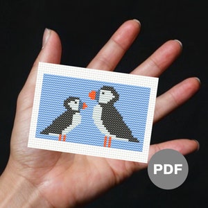 Könnte beinhalten: Ein Kreuzstichmuster mit zwei Papageientauchern auf blauem Hintergrund. Die Papageientaucher schauen sich an und einer hält ein kleines orangefarbenes Herz. Das Muster ist in der unteren rechten Ecke mit "PDF" gekennzeichnet.