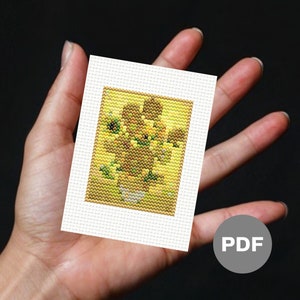 May include: A small, rectangular cross-stitch pattern featuring a yellow and brown sunflower design in a white frame. The artwork is held in a hand against a dark background. A PDF icon is in the lower right corner.