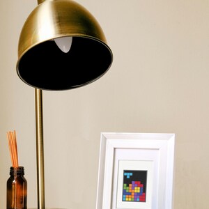 Image of a framed mini cross stitch of an 80s puzzle video game. It is sitting on a table next to a lamp