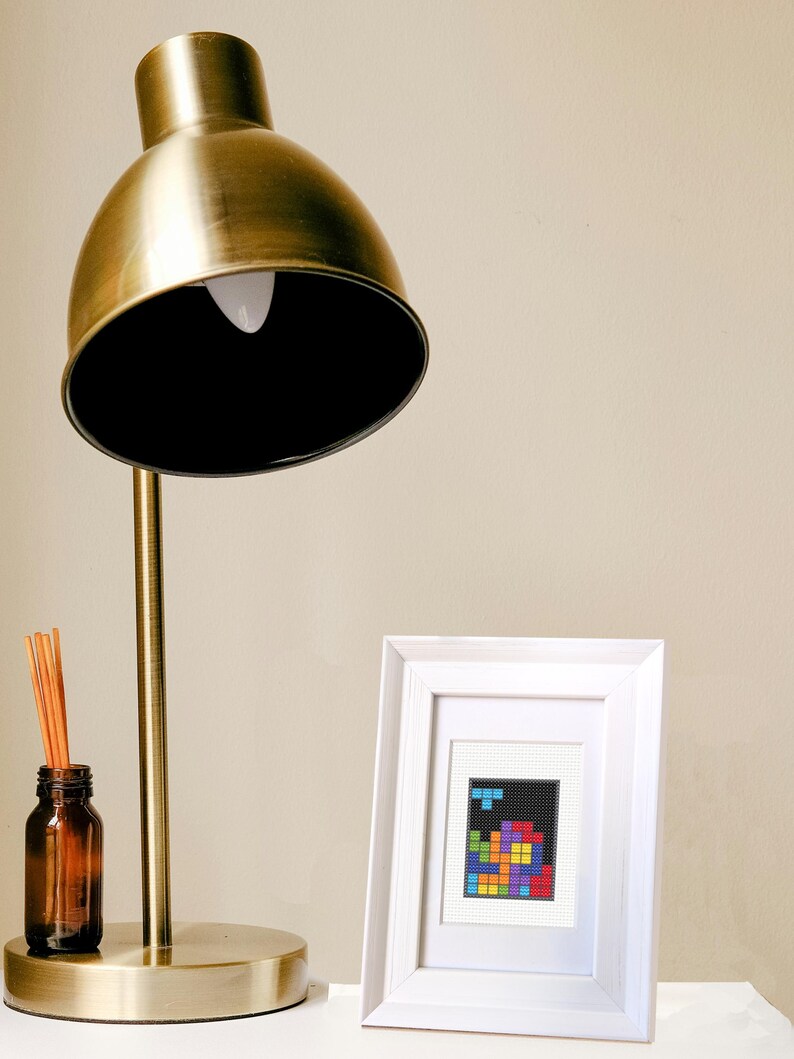 Image of a framed mini cross stitch of an 80s puzzle video game. It is sitting on a table next to a lamp