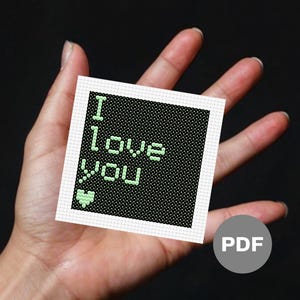 Puede incluir: Un patrón de punto de cruz en blanco y negro con texto verde que dice "Te amo" y un pequeño corazón verde.