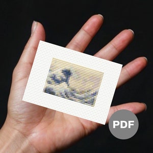 May include: A close-up of a small, white canvas with a cross-stitch pattern of a blue and white wave. The canvas is being held in a hand, and the word "PDF" is printed on a grey circle in the bottom right corner of the image.