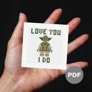 Puede incluir: Un pequeño punto de cruz cuadrado con un personaje verde con el texto "LOVE YOU I DO" en letras verdes. El punto de cruz está sobre un fondo blanco y se sostiene en una mano sobre un fondo oscuro. Un icono PDF está en la esquina inferior derecha.
