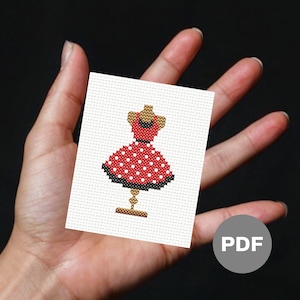 Puede incluir: Un patrón de punto de cruz de un vestido rojo con lunares blancos, un dobladillo negro y un maniquí de color canela. El diseño está sobre un fondo blanco y se sostiene en una mano. Un icono PDF está en la esquina.