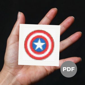 May include: A white canvas with a red, white, and blue embroidered Captain America shield. The shield is in the center of the canvas and is surrounded by a white border. The canvas is being held in a hand. The word "PDF" is written in gray on a gray circle in the lower right corner of the image.