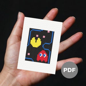 May include: A cross-stitch pattern featuring a red Pac-Man ghost and a yellow Pac-Man pellet on a black background with blue and pink details. The pattern is printed on white paper and labeled "PDF".