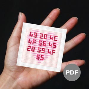 Puede incluir: Un pequeño diseño de punto de cruz cuadrado con forma de corazón rosa con números y letras en un tono rosa más oscuro. El diseño se sostiene en una mano sobre un fondo oscuro. Un icono PDF está en la esquina inferior derecha.