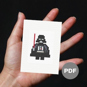 May include: A black and white pixelated cross-stitch pattern of Darth Vader from Star Wars. The pattern is on white fabric and includes a red lightsaber. The image includes the word "PDF" in a gray circle.
