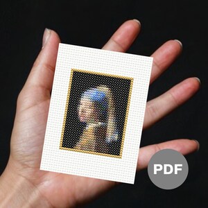 May include: A cross-stitch pattern of the famous Girl with a Pearl Earring painting. The pattern is in a white frame with a gold border. The pattern is for a small cross-stitch project.
