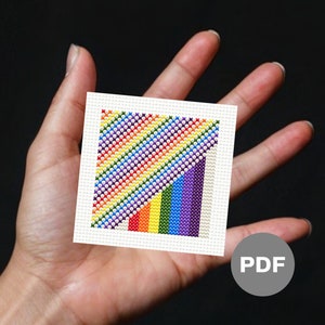 May include: A small, white canvas with a rainbow-coloured diagonal pattern stitched in cross-stitch. The pattern is made up of red, orange, yellow, green, blue, indigo, and violet. The canvas is held in a hand, and a PDF label is visible in the background.