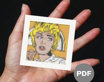 May include: A cross-stitch pattern of a woman with blonde hair, wearing a white shirt and a yellow background. The pattern is in a white frame and is being held in a hand. The image includes the word "PDF" in a gray circle.