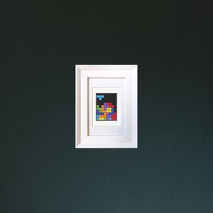 Image of a framed mini cross stitch of an 80s puzzle video game. It is hanging on a dark wall