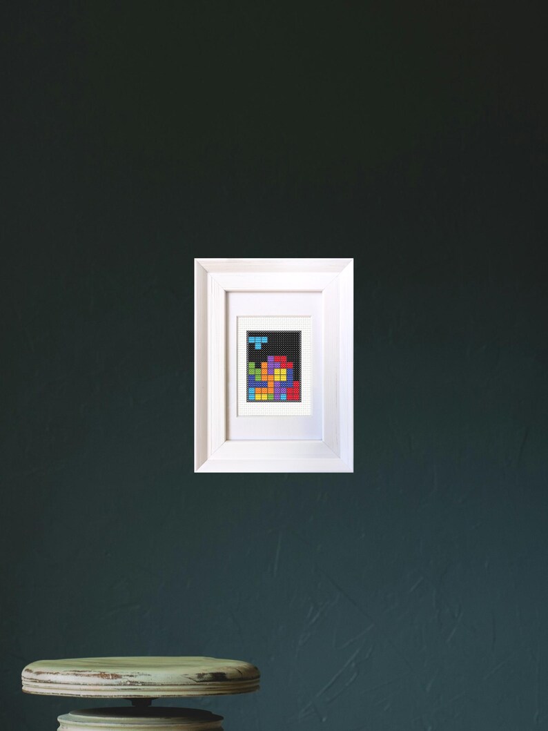 Image of a framed mini cross stitch of an 80s puzzle video game. It is hanging on a dark wall