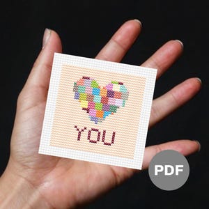 Könnte beinhalten: Ein Kreuzstichmuster mit einem bunten Herzen und dem Wort "YOU" darunter. Das Muster ist auf einer weißen Karte mit hellpfirsichfarbenem Hintergrund gedruckt. Die Karte wird in einer Hand gehalten. Das Bild ist mit "PDF" beschriftet.