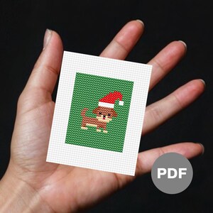 May include: A cross-stitch pattern featuring a brown dog wearing a red Santa hat on a green background. The pattern is on a white card with a PDF label.