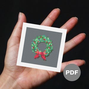 Op de afbeelding: Een digitaal kruissteekpatroon met een groene krans met rode bessen en een rode strik. Het patroon wordt weergegeven op een witte achtergrond met een grijze rand. Het woord "PDF" is in wit gedrukt op een grijze cirkel.