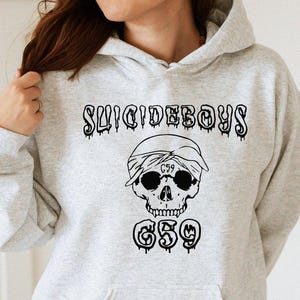 May include: Light gray hoodie with a black graphic featuring the text "SUICIDEBOYS" above a skull wearing a bandana and sunglasses, with "G59" below. The hoodie has a drawstring hood and a front pocket.