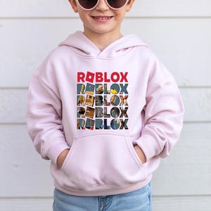K&ouml;nnte beinhalten: Hellrosa Kapuzenpullover mit dem Wort "ROBLOX" in einem gestapelten Design. Das oberste Wort ist rot, die nachfolgenden W&ouml;rter sind mit farbenfrohen Bildern gef&uuml;llt. Der Pullover hat eine Vordertasche und lange &Auml;rmel.