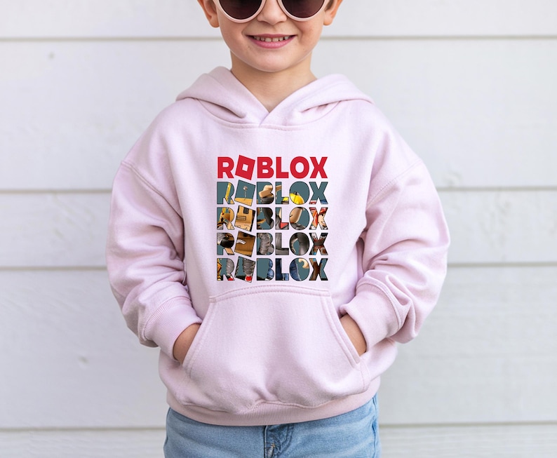 K&ouml;nnte beinhalten: Hellrosa Kapuzenpullover mit dem Wort "ROBLOX" in einem gestapelten Design. Das oberste Wort ist rot, die nachfolgenden W&ouml;rter sind mit farbenfrohen Bildern gef&uuml;llt. Der Pullover hat eine Vordertasche und lange &Auml;rmel.
