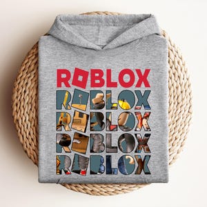 K&ouml;nnte beinhalten: Ein grauer Kapuzenpullover mit dem Wort "ROBLOX" in roten Blockbuchstaben oben. Das Wort "ROBLOX" wird f&uuml;nfmal auf der Vorderseite des Sweatshirts wiederholt, wobei jeder Buchstabe mit einem anderen Bild gef&uuml;llt ist.