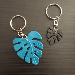 Op de afbeelding: Twee sleutelhangers met een monstera-bladontwerp. De ene sleutelhanger is blauw en de andere is zwart.