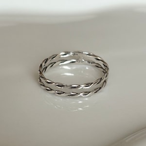 May include: A silver ring with a braided design. The ring features two intertwined bands, creating a textured, woven appearance. The ring is set against a white background, highlighting its metallic sheen. This piece of jewelry is suitable for everyday wear.