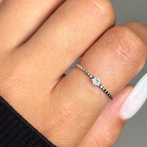 May include: A delicate silver ring with a small, clear gemstone set in the center. The ring is on a finger with a white nail.