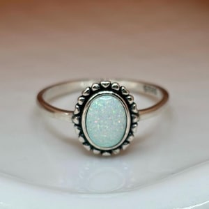 May include: A silver ring with a white opal gemstone set in a decorative oval bezel. The ring has a delicate band and a textured design around the stone.