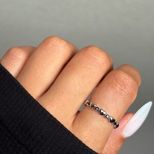 May include: A silver ring with a series of small heart-shaped designs, each with a dark center. The ring is worn on a finger, with a black sleeve visible in the background. The ring is a delicate piece of jewelry.