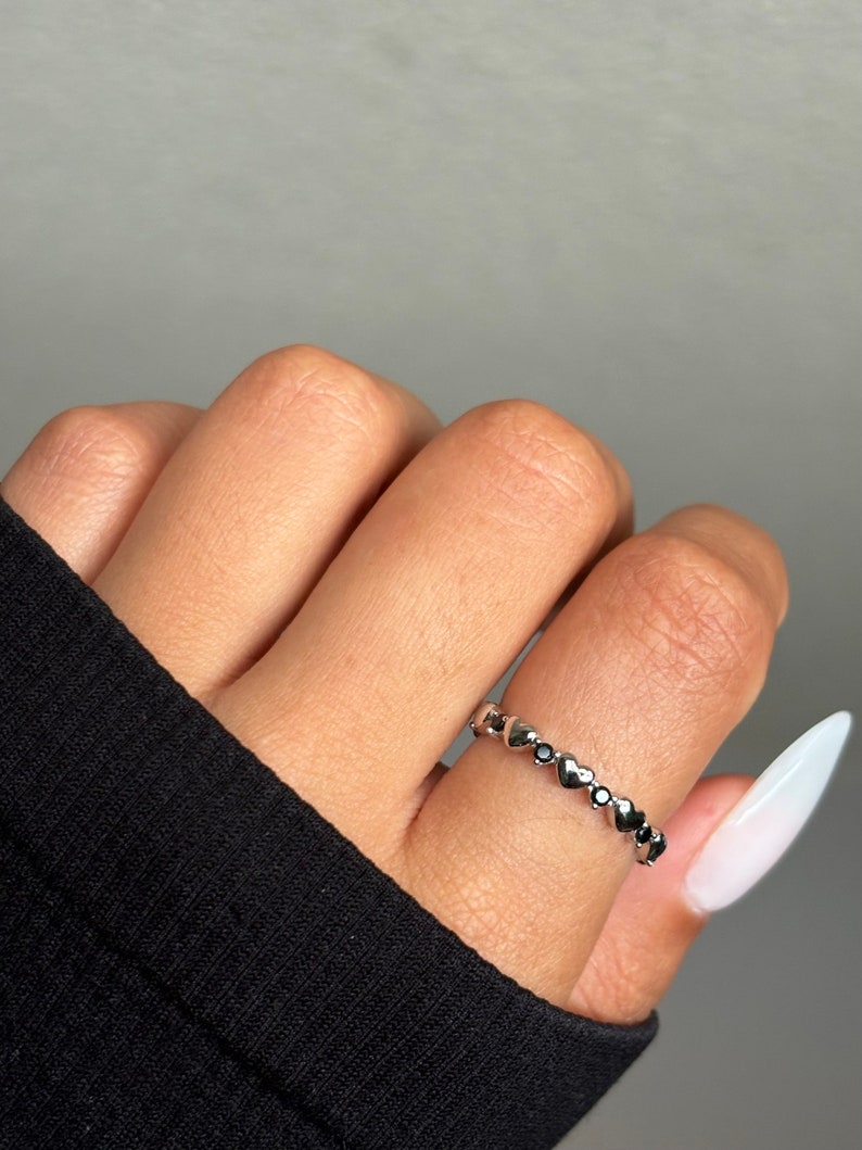 May include: A silver ring with a series of small heart-shaped designs, each with a dark center. The ring is worn on a finger, with a black sleeve visible in the background. The ring is a delicate piece of jewelry.