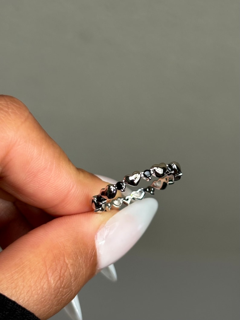 May include: A silver ring featuring a continuous pattern of small hearts. The ring is held between fingers with a light gray background.