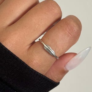 May include: A silver-colored ring featuring a detailed feather design. The ring is worn on a finger, showcasing the intricate lines and texture of the feather. The ring's band is thin and delicate.
