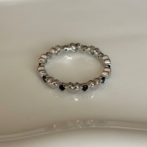 May include: A silver ring featuring a repeating heart design. Small black gemstones are set between the hearts. The ring is displayed on a white surface, showcasing its delicate details and polished finish.