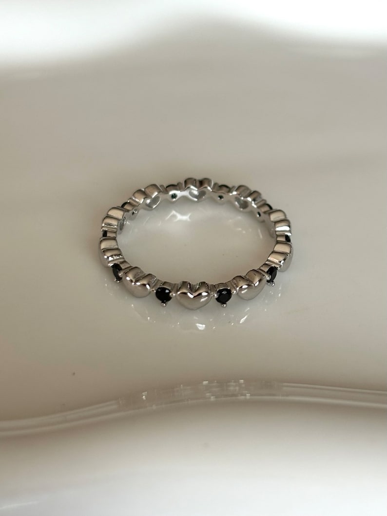 May include: A silver ring featuring a repeating heart design. Small black gemstones are set between the hearts. The ring is displayed on a white surface, showcasing its delicate details and polished finish.