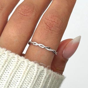 May include: A silver-colored ring with a braided design, adorned with small, sparkling stones. The ring is worn on a finger, showcasing its intricate details and elegant style. The ring is a piece of jewelry.