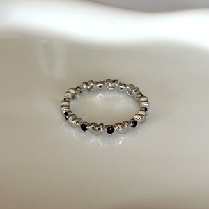 May include: A silver-colored ring featuring a repeating pattern of heart-shaped designs. Each heart is accented with a small, dark-colored stone. The ring is set against a white background, highlighting its delicate details and design.