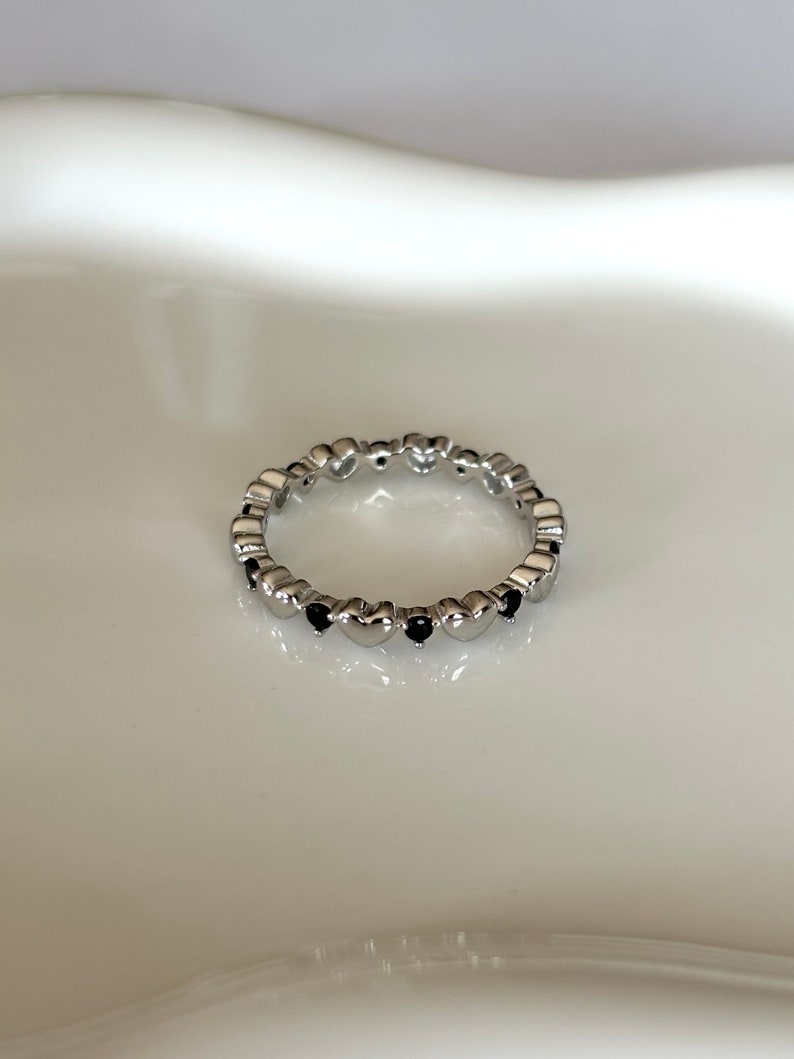 May include: A silver-colored ring featuring a repeating pattern of heart-shaped designs. Each heart is accented with a small, dark-colored stone. The ring is set against a white background, highlighting its delicate details and design.