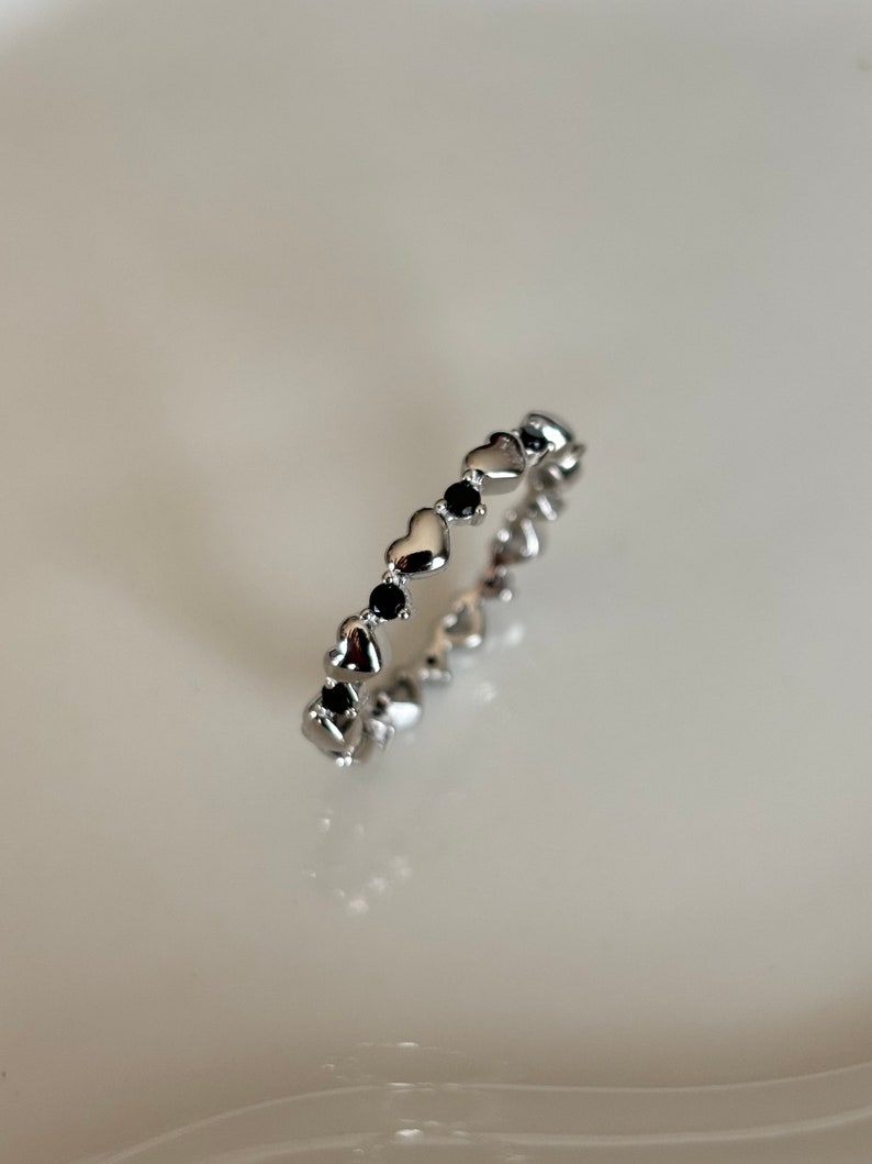 May include: A silver ring featuring a repeating pattern of heart-shaped designs and interspersed with black gemstones. The ring is displayed on a white surface, showcasing its delicate details and reflective qualities. This piece is a romantic and elegant jewelry item.