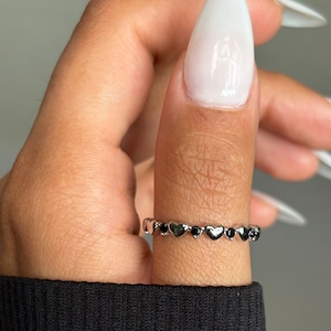 May include: A silver ring with a heart and black stone design is worn on a finger. The ring features alternating heart-shaped and round black stone accents. The person's fingernails are long and painted white.