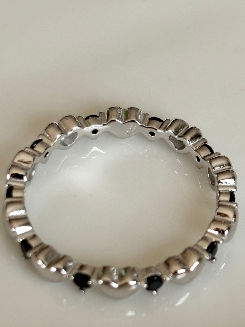 May include: A silver ring with a repeating heart design. Each heart is accented with a small, black gemstone. The ring is made of a shiny, polished metal.