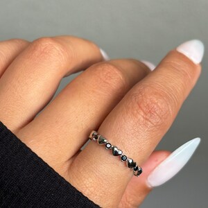 May include: A silver ring featuring a series of heart-shaped designs, interspersed with small black stones. The ring is worn on a finger, showcasing its delicate and romantic aesthetic. The ring is a piece of jewelry.