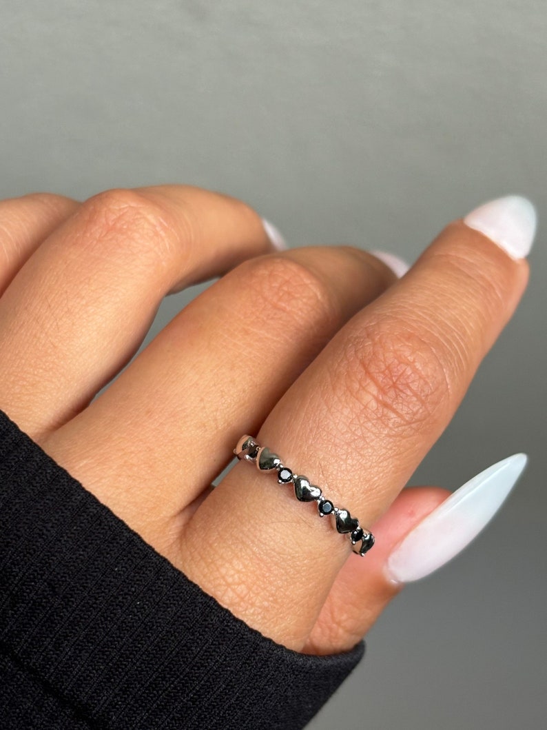 May include: A silver ring featuring a series of heart-shaped designs, interspersed with small black stones. The ring is worn on a finger, showcasing its delicate and romantic aesthetic. The ring is a piece of jewelry.