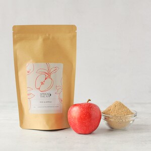 May include: A brown paper bag with a label that says "Spread the Joy" and "Rye & Apple". There is a red apple and a bowl of brown powder in front of the bag.