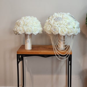 May include: Two white silk flower bouquets with pearl and rhinestone accents on a wooden table with black metal legs.