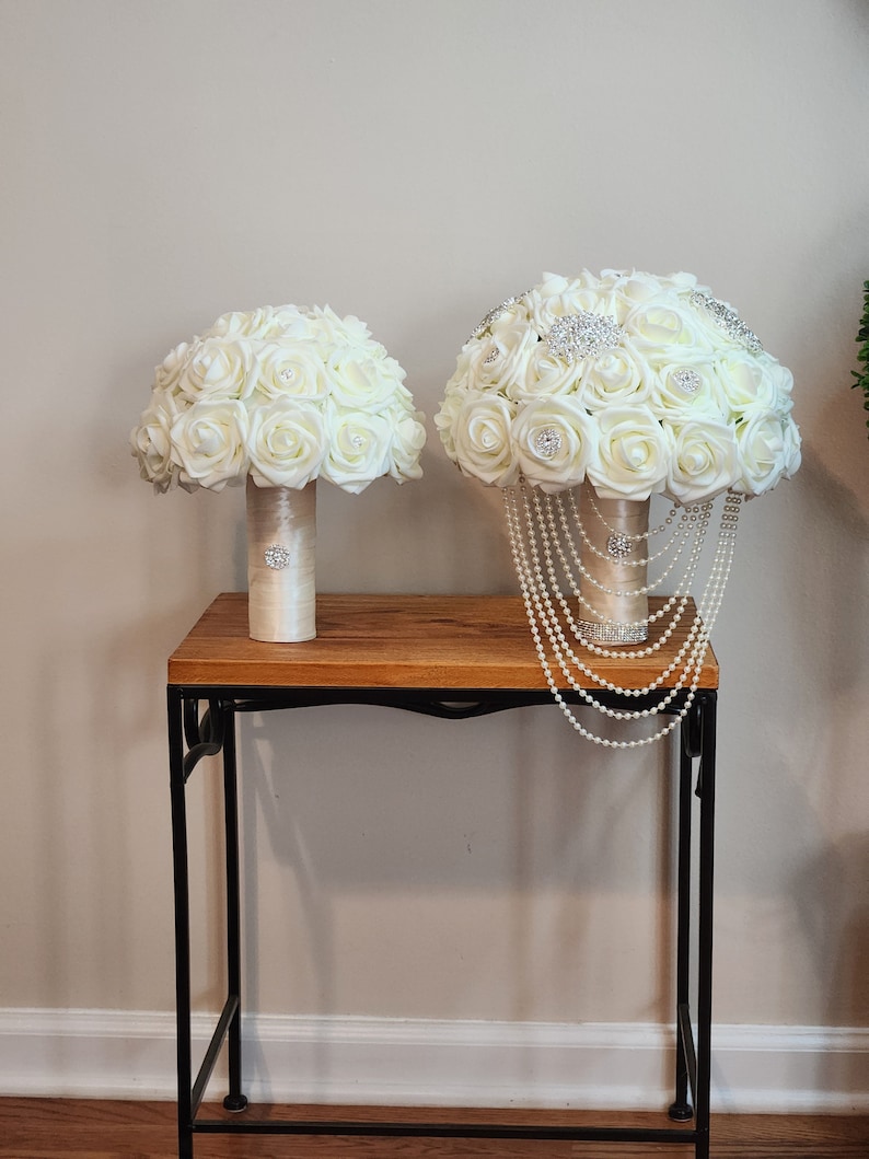 May include: Two white silk flower bouquets with pearl and rhinestone accents on a wooden table with black metal legs.