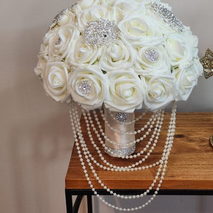 May include: A white bridal bouquet made of artificial roses with pearl accents and rhinestone embellishments. The bouquet is sitting on a wooden table with a pearl garland draped over the side.