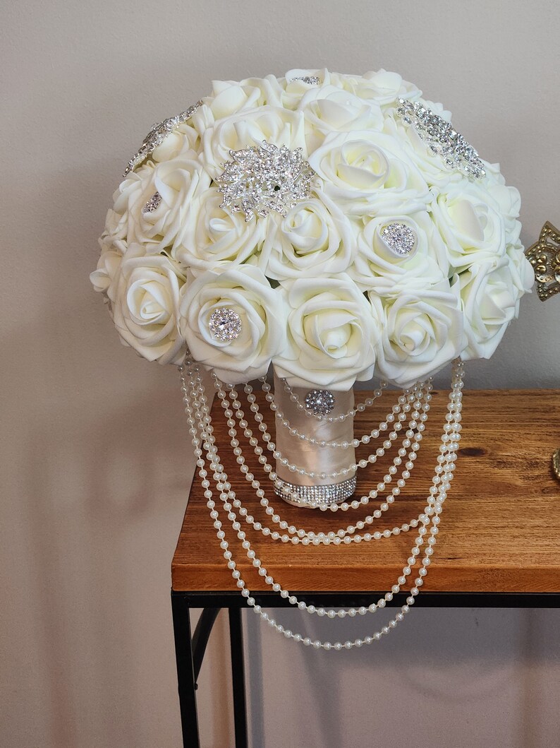 May include: A white bridal bouquet made of artificial roses with pearl accents and rhinestone embellishments. The bouquet is sitting on a wooden table with a pearl garland draped over the side.