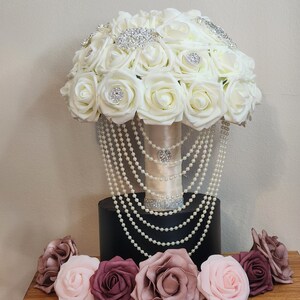 May include: A white bridal bouquet with pearl accents and rhinestone embellishments. The bouquet is surrounded by smaller blush and burgundy roses.